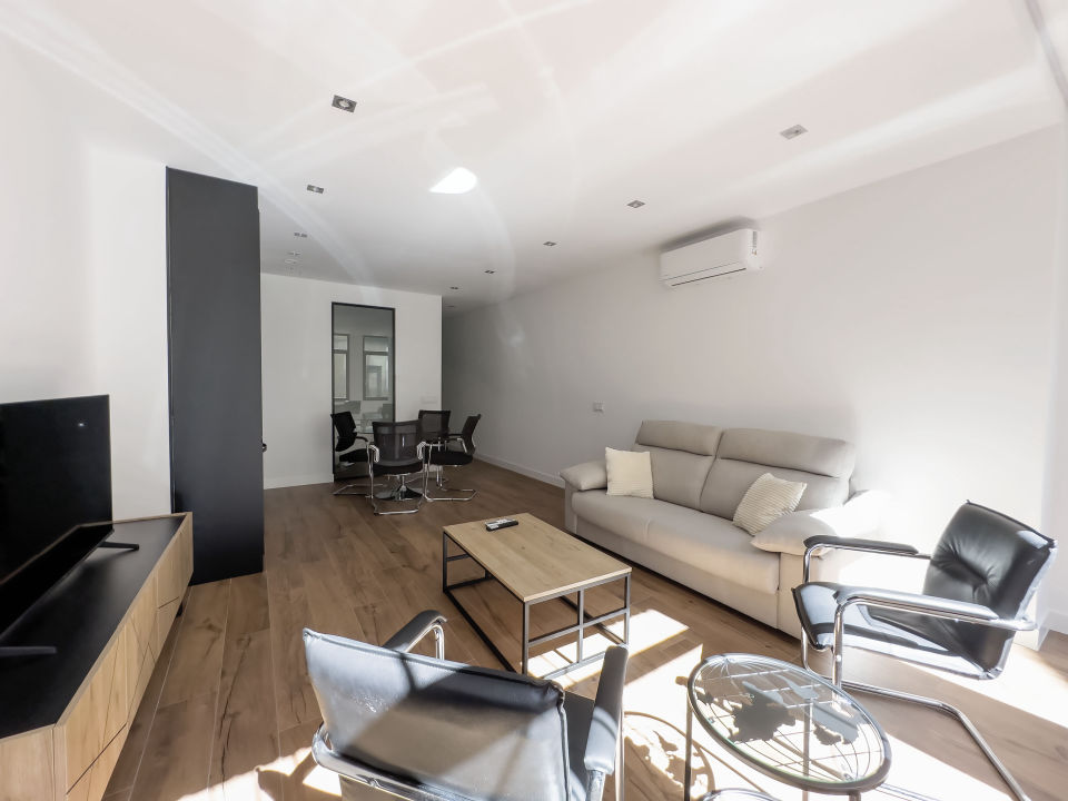 Vista panorámica del apartamento de lujo Alfisa desde el pasillo de entrada, mostrando la zona de estar con sofá, televisión y mueble enfrente. A la derecha, la mesa de comedor con sillas y la cocina en la pared. Al fondo, la zona de estar con mobiliario moderno, sofá cama grande y cómodo.