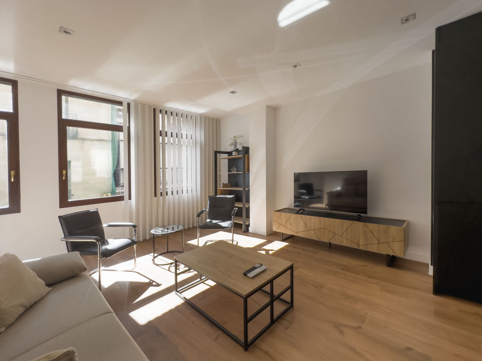 Vista panorámica del apartamento de lujo Alfisa desde el pasillo de entrada, mostrando la zona de estar con sofá, televisión y mueble enfrente. A la derecha, la mesa de comedor con sillas y la cocina en la pared. Al fondo, la zona de estar con mobiliario moderno, sofá cama grande y cómodo.