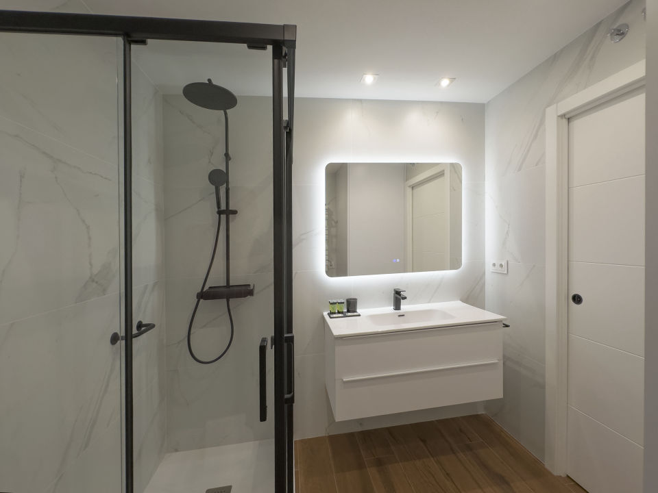 Vista parcial de un baño de lujo en los apartamentos Alfisa, con lavabo de mármol, grifo monomando, ducha con efecto lluvia y mamparas de cristal. Diseño moderno con espejo iluminado y detalles sofisticados