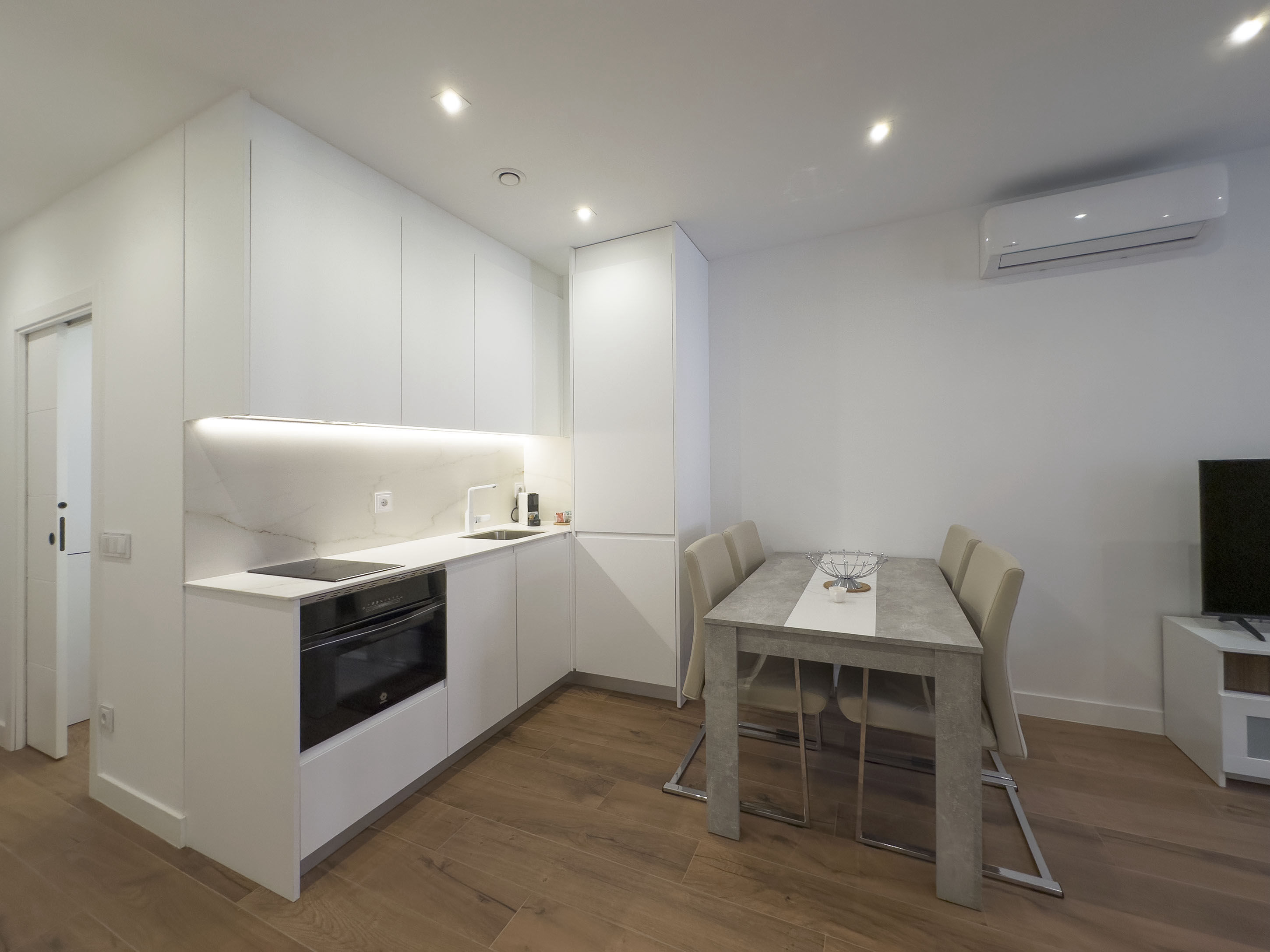 Cocina moderna en los apartamentos de lujo Alfisa, equipada con horno, vitrocerámica, cafetera y mesa de comedor con sillas. La nevera está integrada y oculta. También se aprecia el sistema de aire acondicionado, en un espacio con diseño contemporáneo.