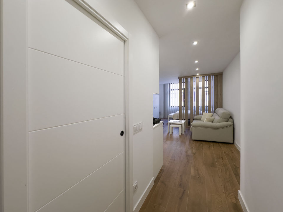 Vista panorámica del apartamento de lujo Alfisa desde el pasillo de entrada, mostrando la zona de estar con sofá, televisión y mueble enfrente. A la izquierda, la mesa de comedor con sillas y la cocina en la pared. Al fondo, un separador de barrotes de madera que divide la cama de matrimonio.