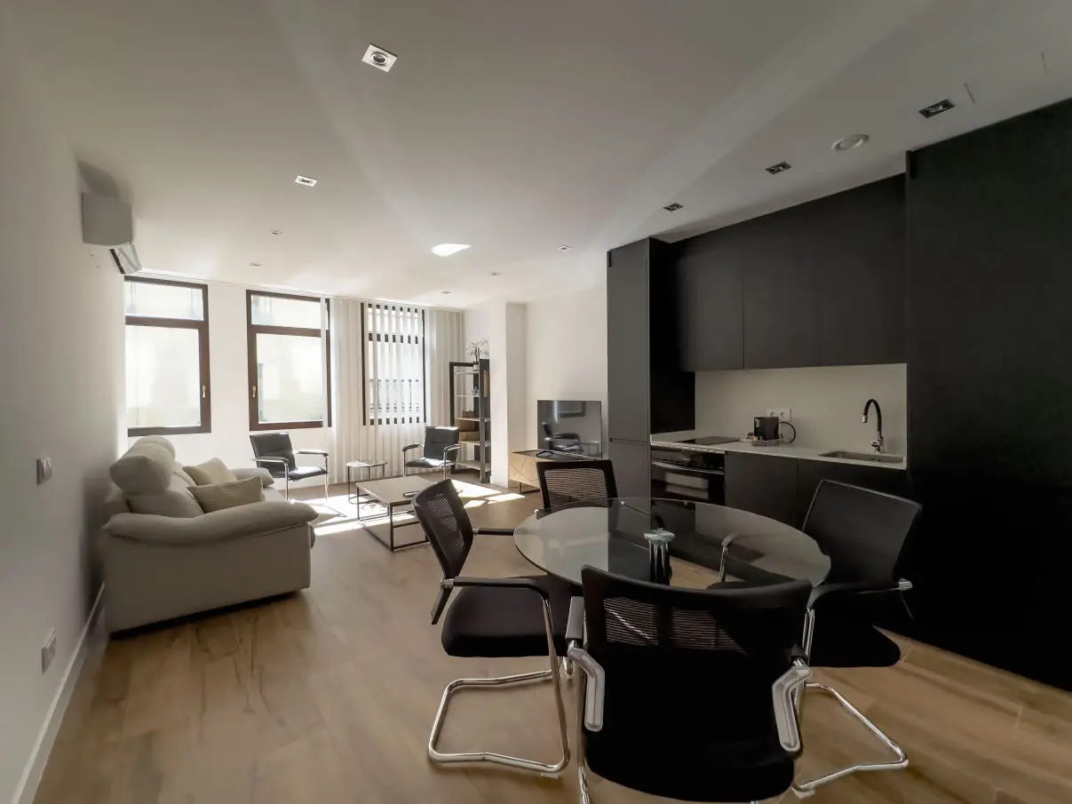 vista general del apartamento de lujo B2 estancia donde en un mismo espacio se sitúan el sofá cama, una mesa comedor con cuatro sillas, un mueble moderno bajo con televisor, y una cocina completa con nevera, horno, vitrocerámica y lavavajillas