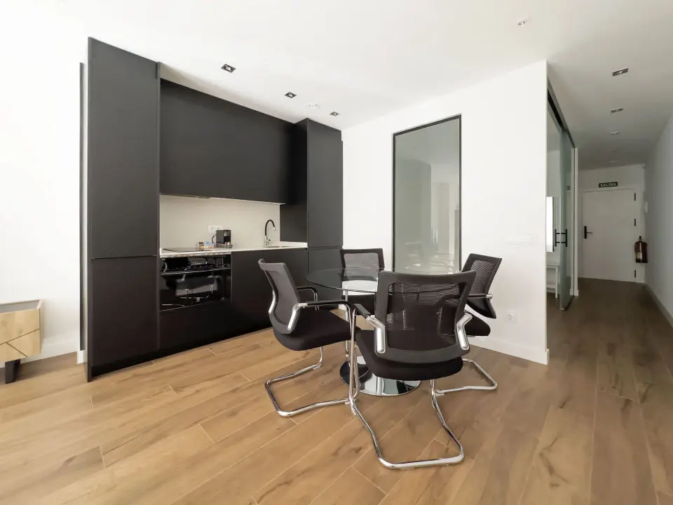 Cocina moderna en los apartamentos de lujo Alfisa, en color negro con la mesa y sillas a juego, equipada con horno, vitrocerámica, cafetera y mesa de comedor con sillas. La nevera está integrada y oculta. También se aprecia el sistema de aire acondicionado, en un espacio con diseño contemporáneo.