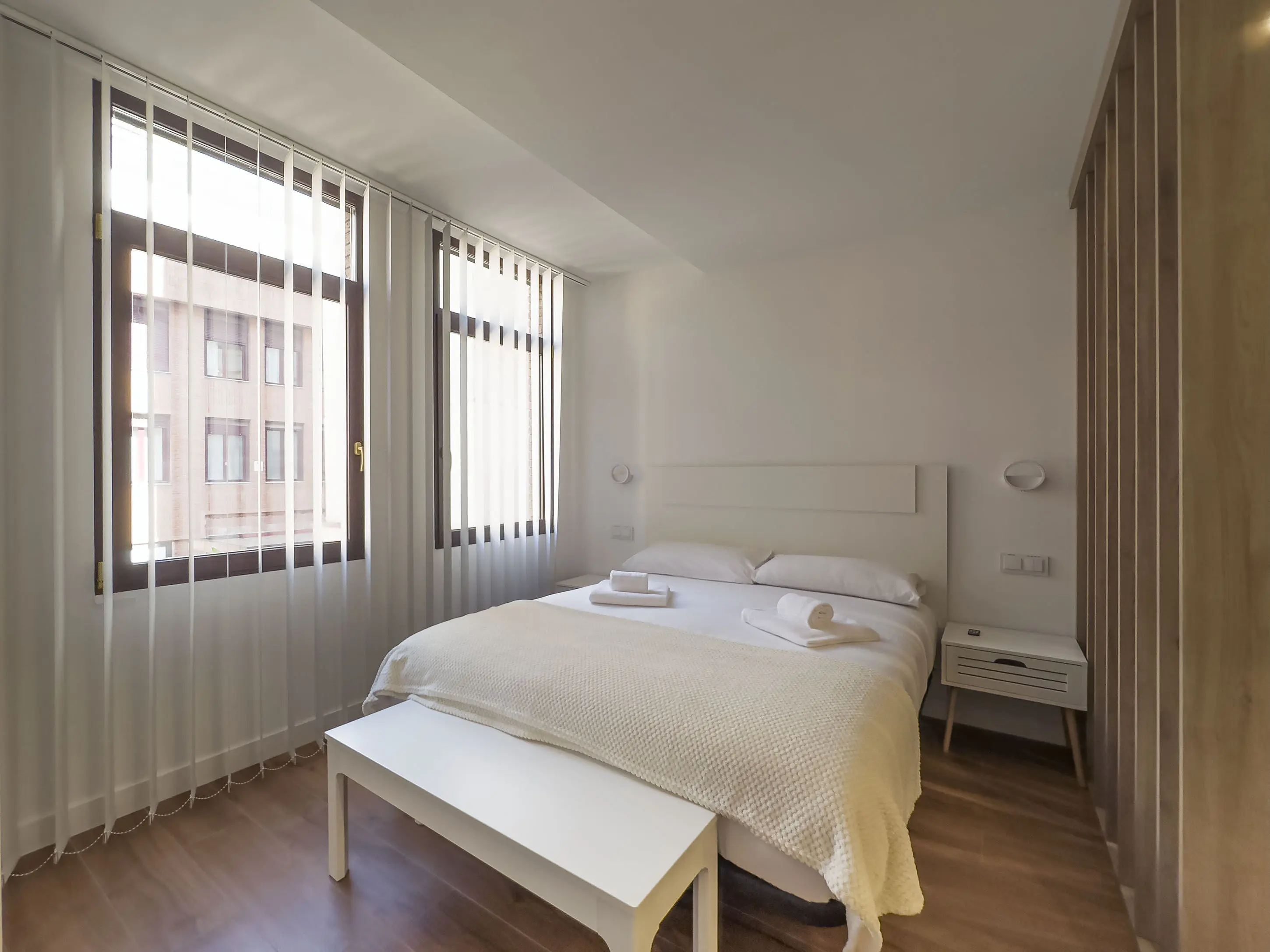 vista general del apartamento desde la habitación de matrimonio en los apartamentos Alfisa, grandes ventanales, mesillas con luces individuales y descalzador de madera blanca a los pies de la cama. Separador de madera artesanal en el lado derecho que divide la estancia del resto del apartamento.