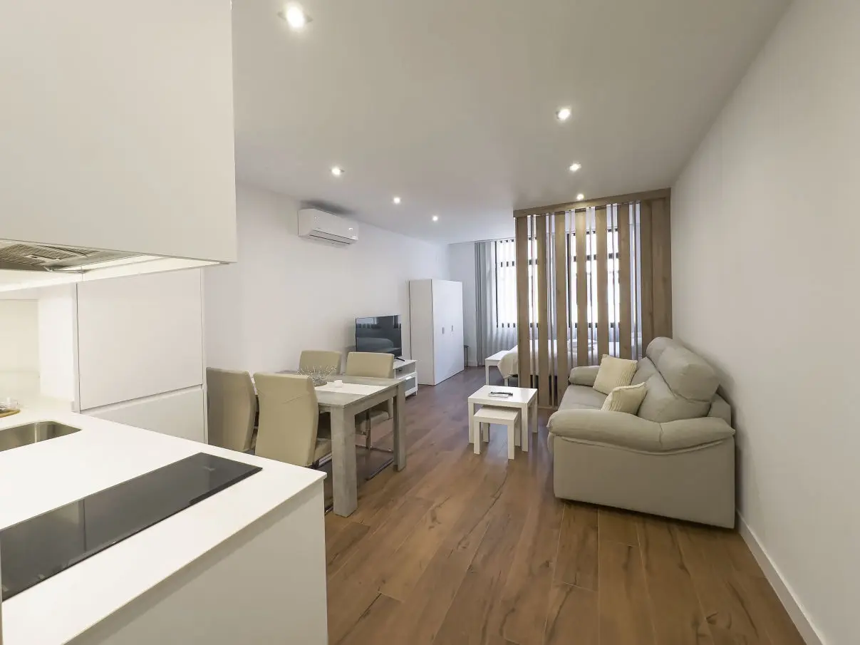 vista general del apartamento de lujo B1 de Alfisa, situado en l calle príncipe 5, a seis minutos a pie de la Puerta del Sol, se ve una cama con separación del resto de la estancia donde en un mismo espacio se sitúan el sofá cama, una mesa comedor con cuatro sillas, un mueble moderno bajo con televisor, y una cocina completa con nevera, horno, vitrocerámica y lavavajillas