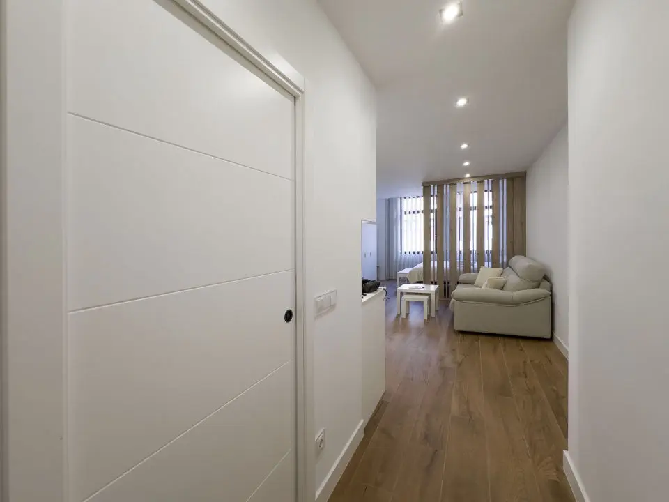 Vista panorámica del apartamento de lujo Alfisa desde el pasillo de entrada, mostrando la zona de estar con sofá, televisión y mueble enfrente. A la izquierda, la mesa de comedor con sillas y la cocina en la pared. Al fondo, un separador de barrotes de madera que divide la cama de matrimonio.