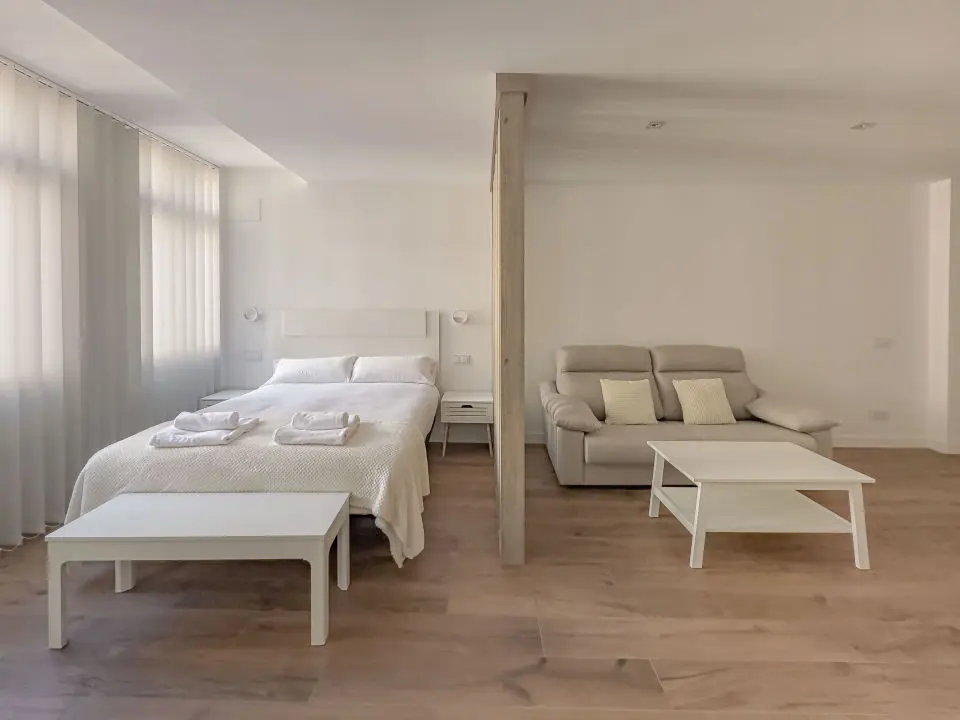 Habitación ultra moderna con cama de matrimonio en los apartamentos Alfisa, separada con grandes cristaleras traslúcidas del resto de las instancias, mesillas con luces individuales y armario de madera blanca a los pies de la cama.