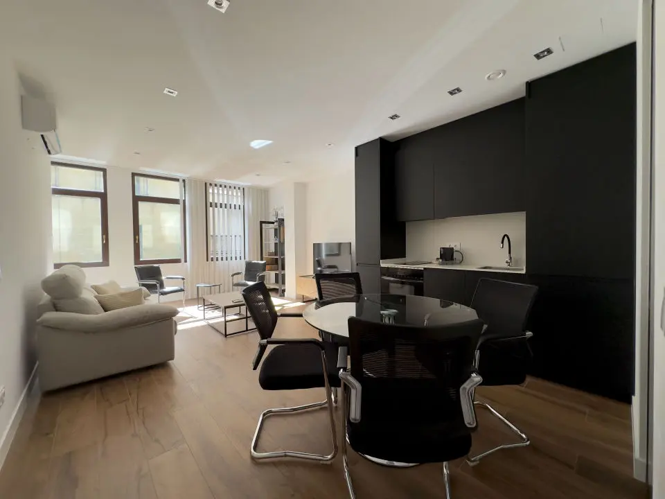 Habitación ultra moderna con cama de matrimonio en los apartamentos Alfisa, separada con grandes cristaleras traslúcidas del resto de las instancias, mesillas con luces individuales y armario de madera blanca al lado de la cama.